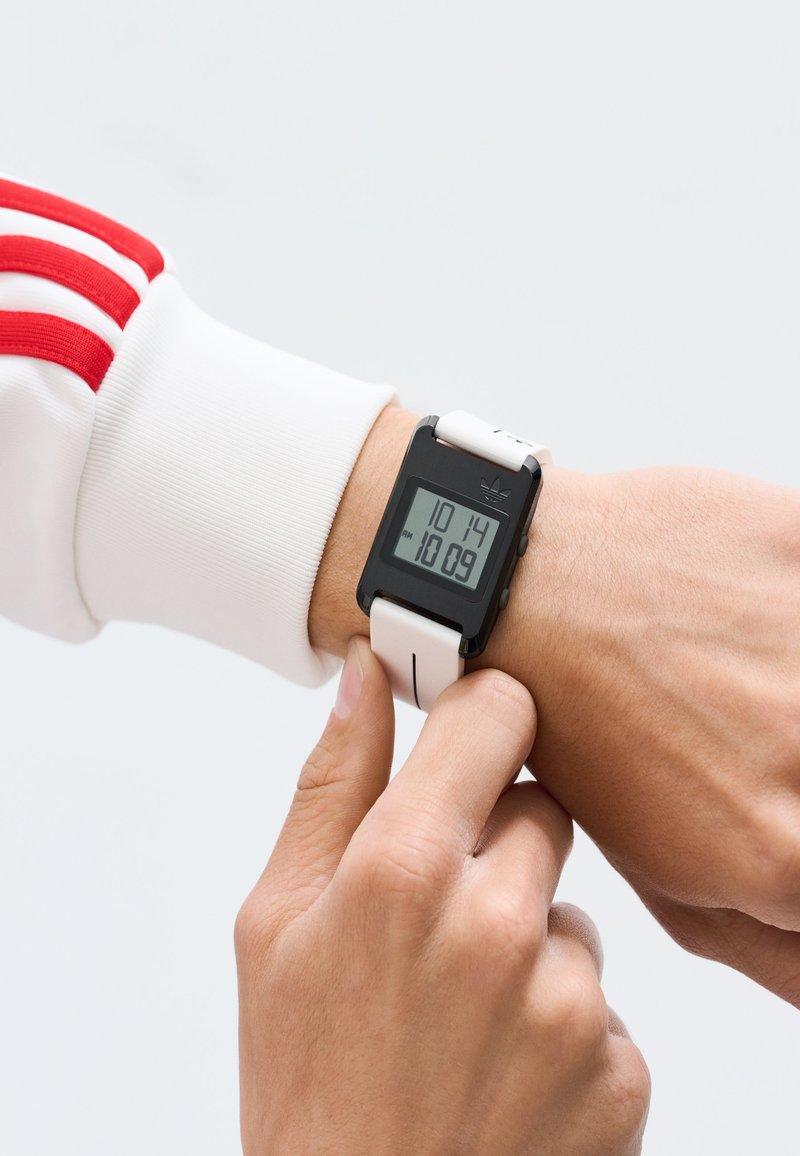 Asmuo, vilkintis baltas ir raudonas rankoves, reguliuoja juodai baltą skaitmeninį laikrodį, rodantį 10:14, šviesiame fone.