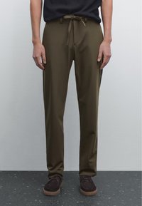 Pantalons légers de couleur vert olive avec un cordon de serrage à la taille, présentant une coupe droite et une texture lisse, associés à des chaussures marron foncé.