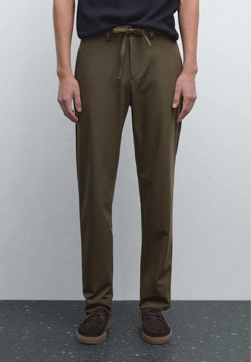 Pantalons légers de couleur vert olive avec un cordon de serrage à la taille, présentant une coupe droite et une texture lisse, associés à des chaussures marron foncé.