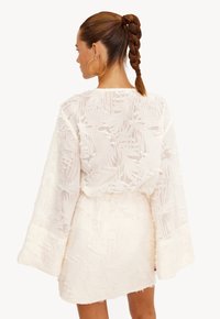 Witte jurk van textuurstof met een palmbladpatroon, lange klokmouwen en een aansluitende taille. De achterkant heeft een ronde hals.