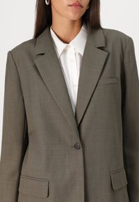 Blazer marron ajusté avec une texture lisse, revers crantés, fermeture à un bouton et poches latérales. Porté sur une chemise blanche à col.