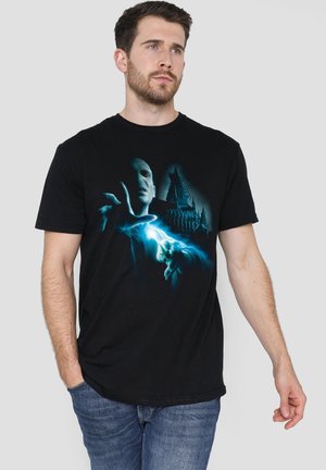 Mann trägt schwarzes T-Shirt mit blauer Grafik einer verhüllten Gestalt, die vor einer dunklen Burg einen Zauber wirkt, vor einfarbigem Hintergrund.