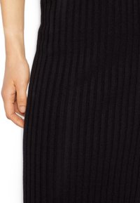 Schwarzer gerippter Strickstoff mit vertikalen Linien, auf dem eine Hand an der Seite ruht. Die Textur scheint dehnbar und weich zu sein.