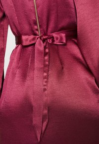 Robe en satin rose fuchsia sur mesure avec manches longues, fermeture éclair dorée à l'avant et détail de nœud à la taille.