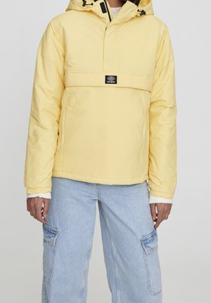 Personne portant une doudoune à capuche jaune clair et un jean cargo bleu clair, debout devant un fond uni.