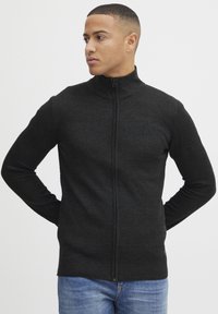 Svart zip-up tröja med hög krage, strukturerad stickad mönster och ribbade ärmslut. Bärs med blåjeans, modellen står med händerna bakom ryggen.