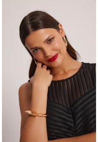 Czarny top o teksturowanej powierzchni z pionowymi paskami z siateczki, zdobiony wyrazistą złotą bransoletą na nadgarstek. Modelka ma długie włosy i wyrazisty czerwony makijaż ust.