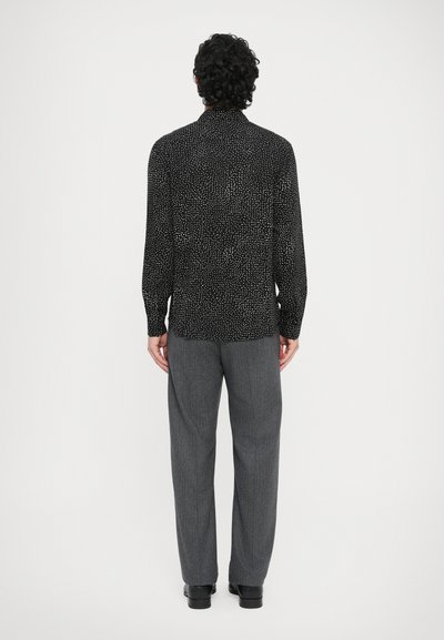 Schwarzes Langarmshirt mit dezentem weißem Punktemuster, kombiniert mit grauen Hose mit weitem Bein und schwarzen Schuhen, von hinten gesehen.