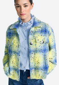 Večbarvna tvidi jakna v rumeni in modri barvi, z teksturirano površino, dvema sprednjima žepoma in srebrnimi gumbi, nošena čez modro črtasto bluzo.