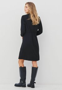 Černé šaty s dlouhým rukávem a elastickým pasem, s dvěma bočními kapsami, doplněné černými koženými botami ke kolenům. Hladký materiál a jednoduchý design.