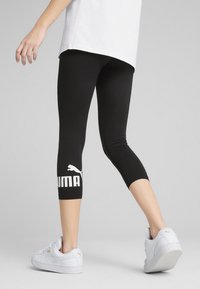 Leggings noirs courts en tissu extensible, avec un logo Puma blanc sur la jambe inférieure. Associés à des baskets blanches.