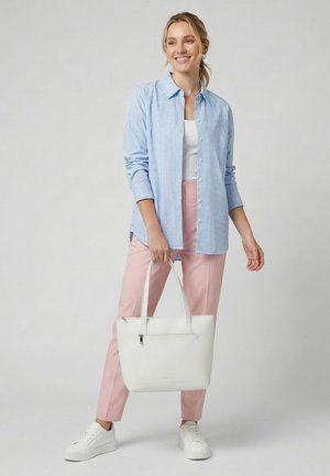 Mujer con camisa de cuadros azul claro, camiseta blanca, pantalones rosas, zapatillas blancas sosteniendo una bolsa tote blanca, sonriendo sobre un fondo neutro.