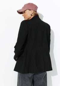 Blazer noir à rayures avec une coupe ajustée. Porté avec une casquette rose clair et des boucles d'oreilles créoles. La vue arrière montre un ourlet légèrement évasé et des poignets boutonnés.
