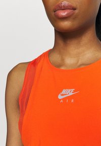 Pomarańczowy top na ramiączkach z siateczkowym wykończeniem na ramionach, z srebrnym logo Nike Air z przodu. Gładka tekstura i sportowy krój.