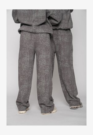 Două persoane purtând pantaloni largi de culoare gri, texturați, și bluze asortate, stând una lângă cealaltă pe un fundal simplu.