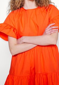 Personne avec des cheveux bouclés portant une robe orange vif avec des manches fronc ées, les bras croisés sur la poitrine, sur un fond blanc uni.