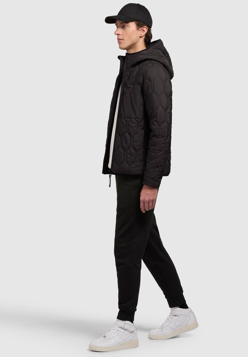 Veste matelassée noire avec capuche, dotée d'un motif hexagonal, associée à un pantalon de jogging noir et des baskets montantes blanches.