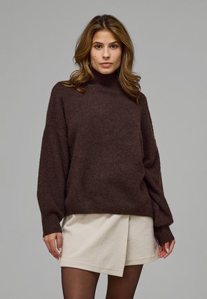 Mujer joven con cabello castaño suelto que lleva un jersey de cuello alto marrón oscuro y una falda cruzada beige con medias negras.