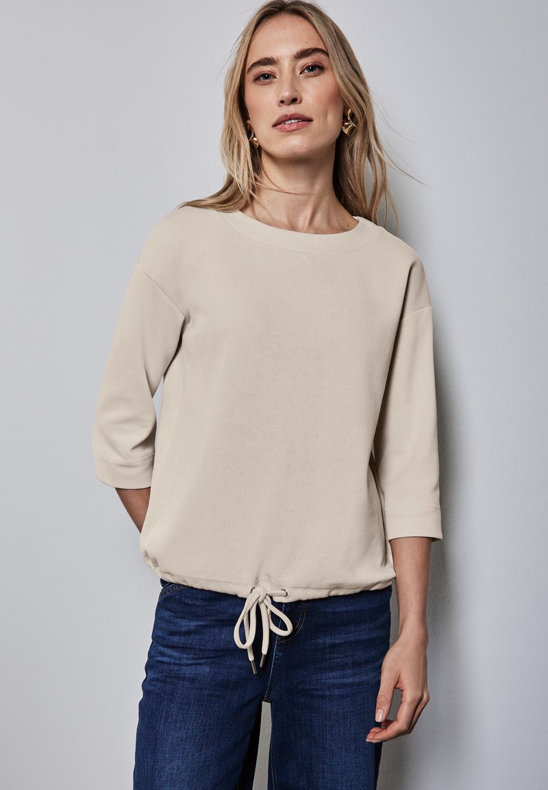 Béžová blůza s kulatým výstřihem, tříčtvrtečními rukávy, stahovací šňůrkou v pase a měkkou texturou, doplněná tmavě modrými džíny.