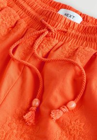 Helle orangefarbene Hosen mit elastischem Bund, bestickt mit Blumenmustern, und Kordeln mit Perlen und Quasten am Ende.