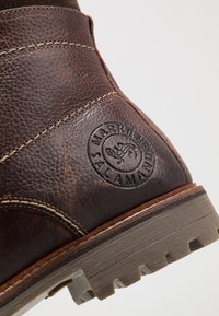 Tacón de bota de cuero marrón texturizado con costuras visibles, logo circular de la marca Salamandra grabado, y suela de goma gruesa.