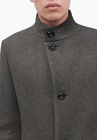 Homme portant un manteau en laine gris avec un col montant et trois boutons noirs visibles, lèvres fermées, sur fond blanc.
