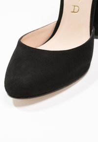 Chaussure à talon bas en daim noir, bout amande, doublure intérieure beige avec logo doré partiellement visible.