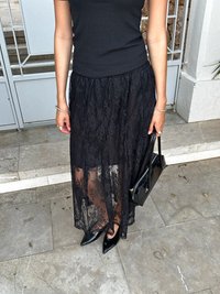 Black lace skirt with a sheer overlay, paired with a fitted black top. Black pointed-toe shoes and a small black handbag complete the look.