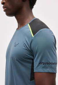 Κοντινό πλάνο ενός ατόμου που φοράει μπλε αθλητικό πουκάμισο με μαύρες πινελιές στους ώμους, κίτρινη λωρίδα και το λογότυπο της Dynafit στο στήθος και το μανίκι.