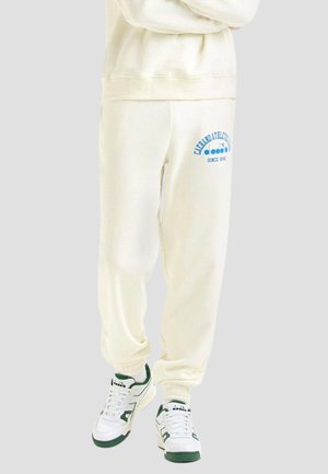 Pantaloni della tuta bianchi in pile con testo stampato blu, polsini a coste e una vestibilità rilassata, abbinati a sneakers bianche con accenti verdi.