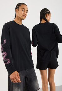 T-shirt noir à manches longues avec des graphiques de serpent rose sur les manches. Design à col ras du cou, fabriqué dans un matériau doux avec une coupe décontractée.