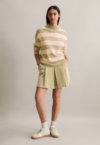 Hellrosa und grün gestreifter Strickpullover mit hohem Kragen; kombiniert mit einem plissierten, hellgrünen Cordrock und beigen Sneakers.