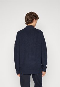 Pull en tricot bleu marine avec col montant, poignets et ourlet côtelés, présentant un tissage texturé et des détails subtils sur les épaules.