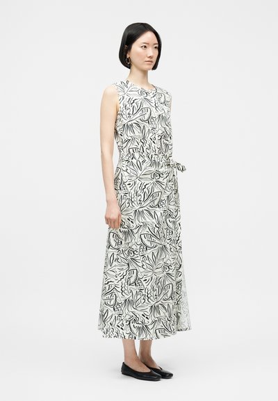 Γυναίκα που φοράει λευκό φόρεμα χωρίς μανίκια, μήκους μέχρι τους αστραγάλους, με μαύρα σχέδια φύλλων και λουλουδιών, δεμένο στη μέση, συνδυασμένο με μαύρα επίπεδα παπούτσια.