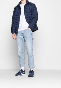 Person klädd i en marinblå puffjacka, vit t-shirt, ljusblå jeans, vita strumpor och marinblå sneakers som står mot en neutral bakgrund.