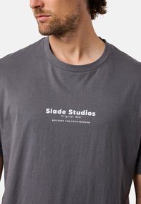 Harmaa puuvillapaita, jossa on pyöreä kaula-aukko ja lyhyet hihat, valkoisella printatut tekstit: "Slade Studios Original Wear Designed for Your Freedom."