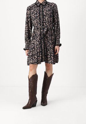 Femme portant une robe noire à fleurs à manches longues avec une ceinture à la taille et des bottes de cowboy brodées brun foncé, montant jusqu'aux genoux.