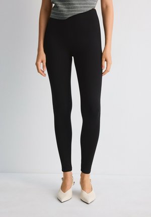 Femme portant un legging noir près du corps avec un top gris à rayures et des talons slingback beiges à bout pointu, debout devant un fond uni.