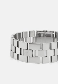 Bracelet en métal argenté avec des maillons rectangulaires, présentant une finition polie et un logo gravé. Fermeture sécurisée visible ; design géométrique moderne.