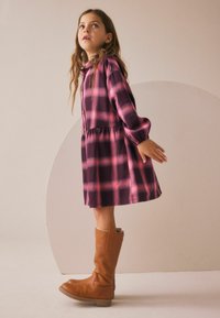 Abito a quadri viola e rosa con maniche lunghe e vita arricciata, abbinato a stivali marroni sopra il ginocchio. Design semplice, tessuto morbido.