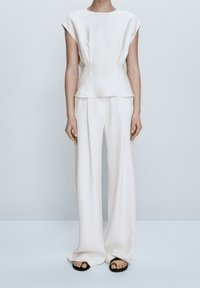 Blouse blanche à manches courtes avec un design structuré, associée à un pantalon à jambes larges. Tissu fluide avec un léger éclat, complété par des sandales simples.