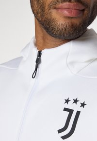 Hvid zip-up jakke med høj krave, sort logo og tre stjerner, lavet af glat stof med en tekstureret finish.