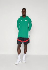 Zelena Nike jopa z logotipom, črne kratke hlače z rdečimi poudarki in beli športni copati. Model stoji pokončno z rokami ob straneh.