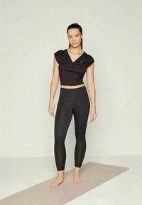 Donna in piedi a piedi nudi su un tappetino yoga beige, indossando un top corto nero e leggings neri, su uno sfondo chiaro e uniforme.