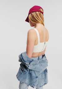 Witte cropped top met textielstructuur en brede banden. Gecombineerd met een lichtdenim jack en een bordeauxrode baseballcap. Achteraanzicht weergegeven.
