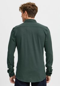 Donkergroen overhemd met lange mouwen, een kraag, en knoopsluiting bij de manchet. De achterkant heeft een gebogen zoom en een eenvoudig ontwerp.