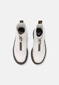 Botas de tobillo blancas de cuero suave, con una cremallera frontal, suela negra con costuras amarillas y un tirador amarillo en la parte trasera.