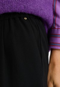 Pantalones negros de corte relajado con cintura fruncida, que incluyen un pequeño botón decorativo dorado. Tejido de textura suave bajo un suéter morado.