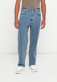 Άντρας που φοράει ανοιχτό γαλάζιο παντελόνι με ίσια γραμμή και λευκά αθλητικά παπούτσια, στέκεται μπροστά σε μια λευκή φόντο.