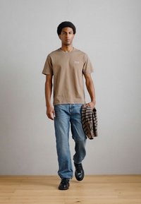 Világosbarna, rövid ujjú póló, kicsi zsebbel, kék farmernadrág és fekete cipő. Tart egy barna csíkozott pulóvert. Fa padló háttér.